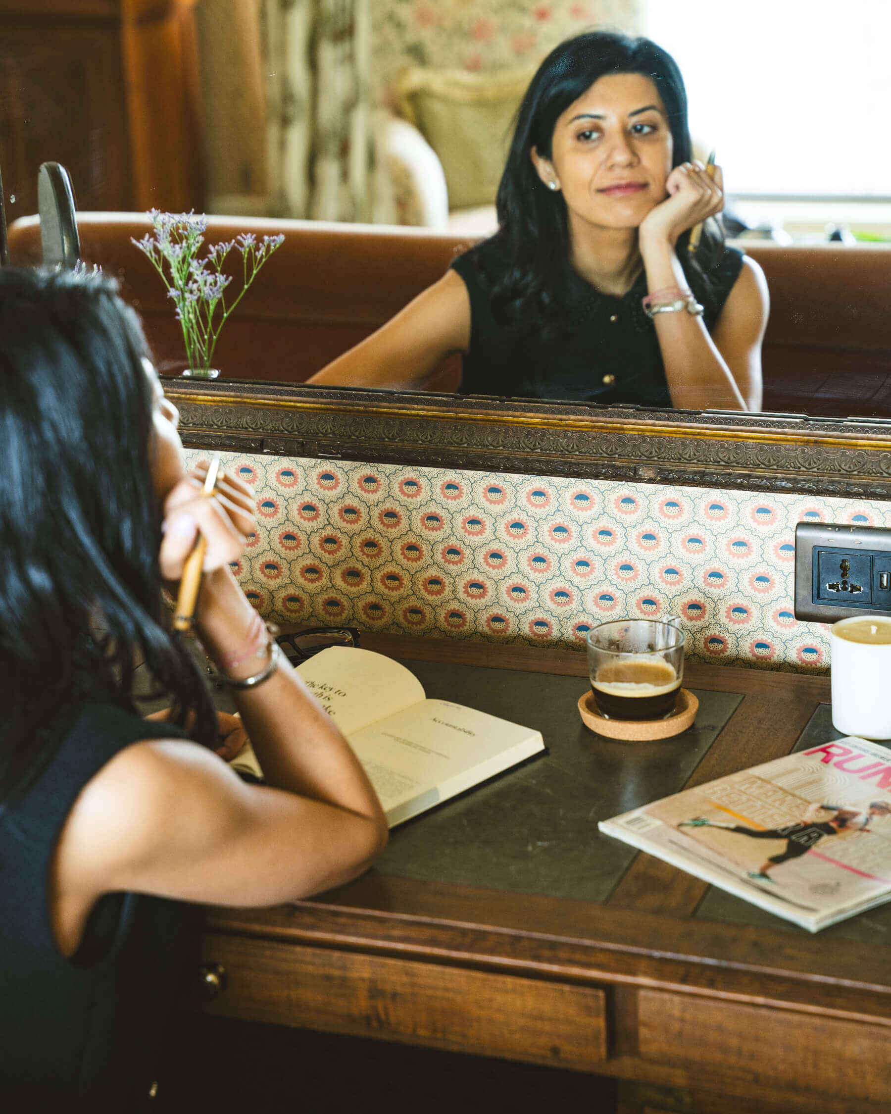 Nikhita, wellness writer, thoughtful and smiling, sitting at a desk in front of a mirror, holding a pen and book on the desk.